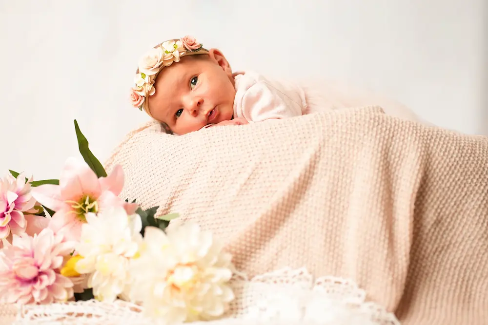 Baby mit Blumenstirnband schaut über eine hellrosa Strickdecke in die Kamera