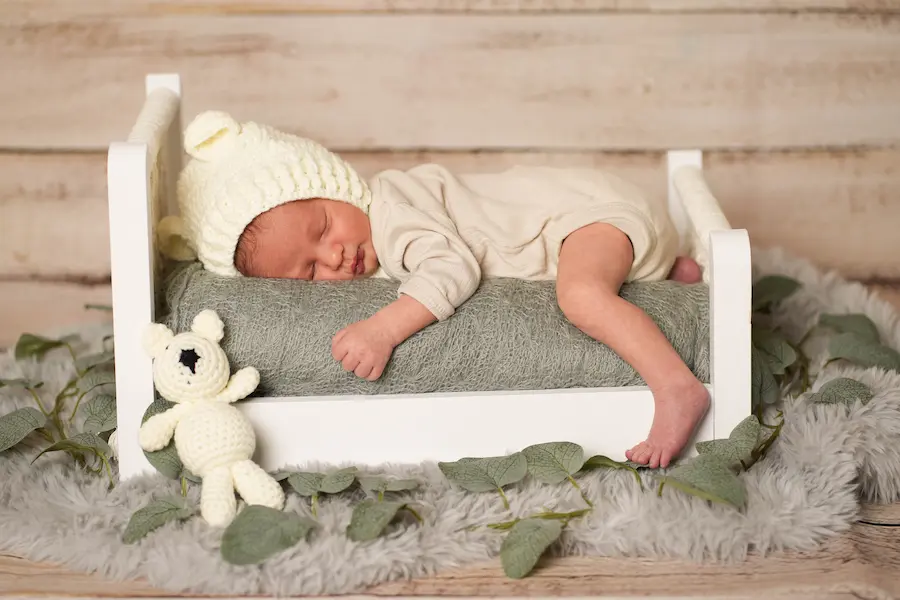 Baby mit weißer Strickmütze schläft quer in einem kleinen Holzbett neben einem Häkelbären