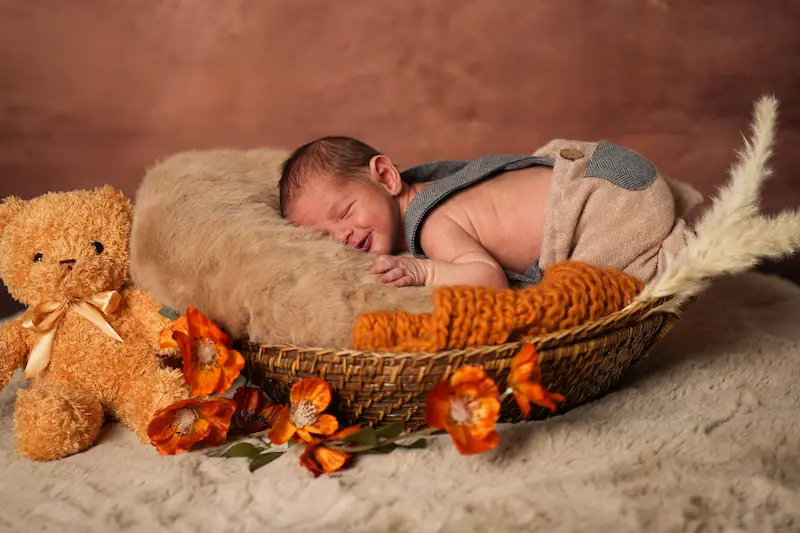Schlafendes Baby mit Hosenträgern im Korb neben einem Teddy und orangefarbenen Blüten