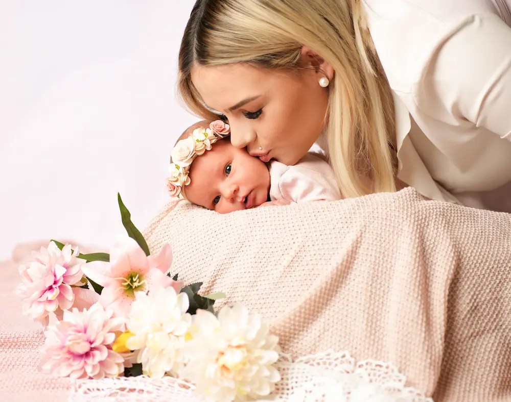 Frau küsst ein waches Baby mit Blumenstirnband, dessen Kopf auf einer rosa Decke ruht