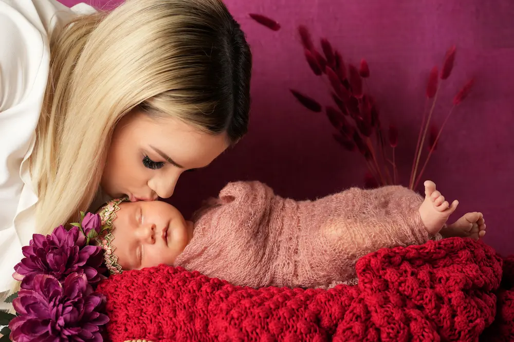 Frau beugt sich für einen Kuss über ein eingewickeltes Baby auf roter Decke vor violettem Hintergrund