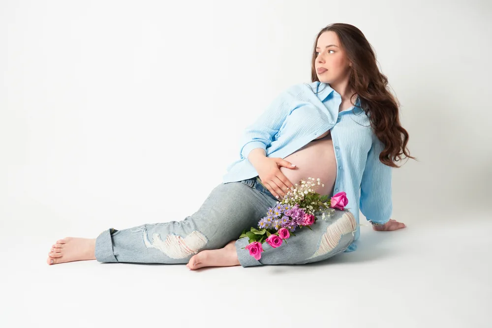 Schwangere Frau im offenen blauen Hemd sitzt seitlich auf dem Boden; rosa Blumen liegen auf ihren Jeans