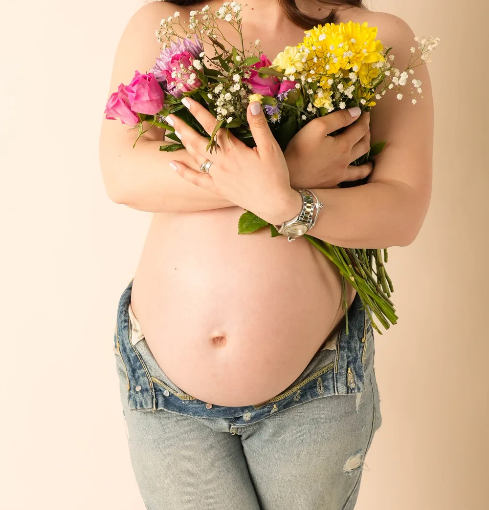 Nahaufnahme vom Babybauch über dem Jeansbund und einem bunten Blumenstrauß in verschränkten Armen