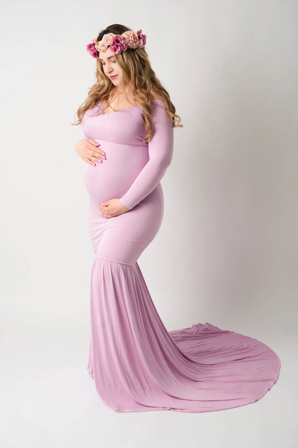 Schwangere Frau im bodenlangen fliederfarbenen Kleid mit Blumenkrone und Händen am Babybauch