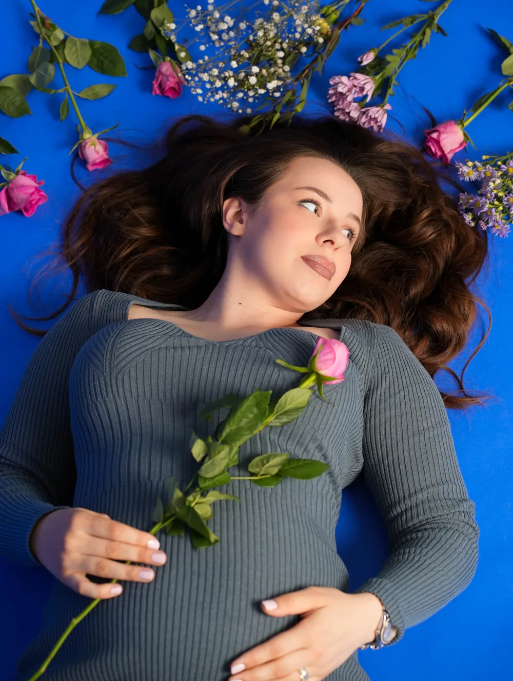 Aufsicht auf eine schwangere Frau im grauen Kleid mit rosa Rose in der Hand und Blumen um ihr Haar