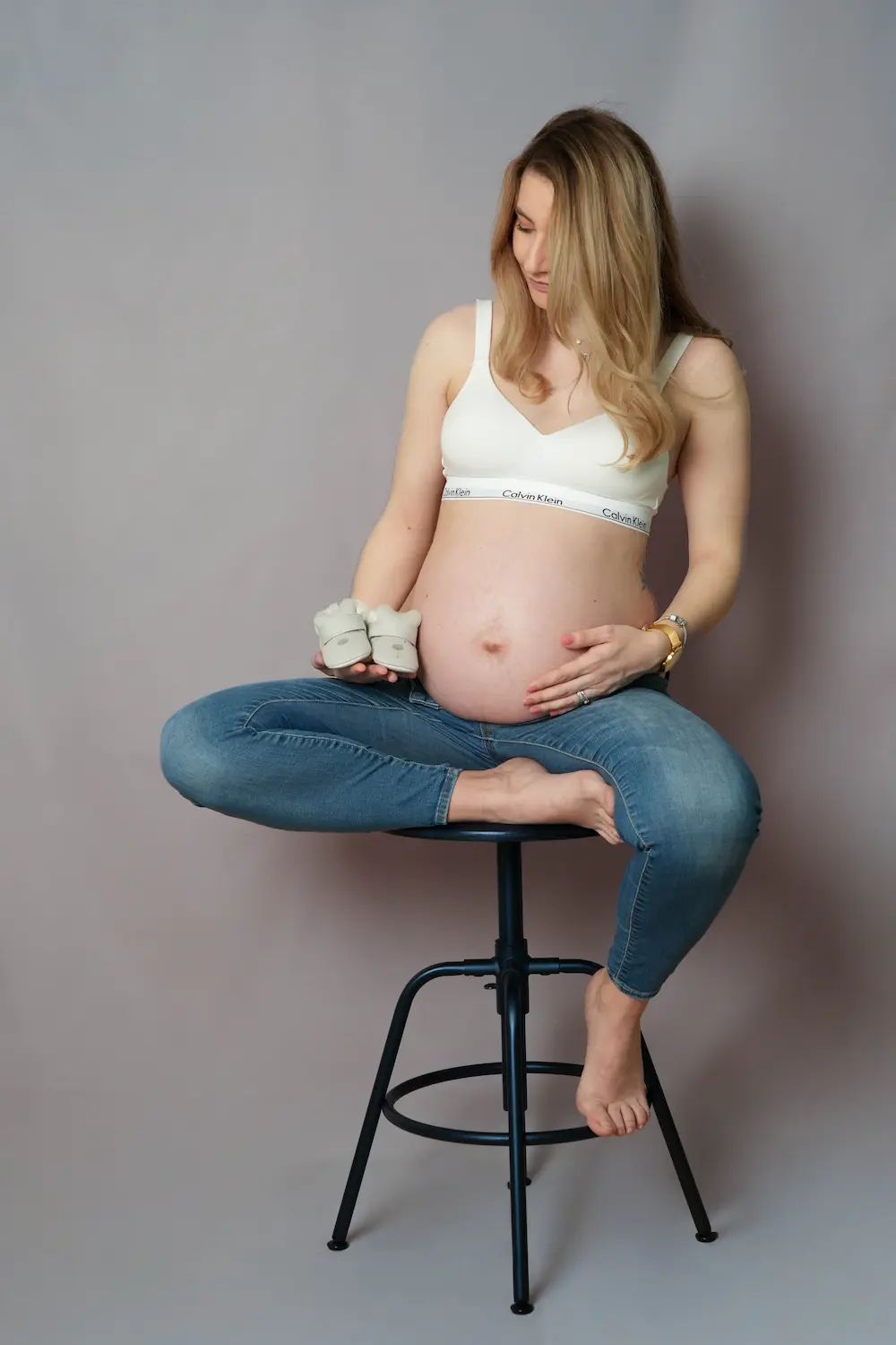 Schwangere Frau sitzt im weißen Oberteil und in Jeans auf einem Hocker und hält kleine Babyschuhe