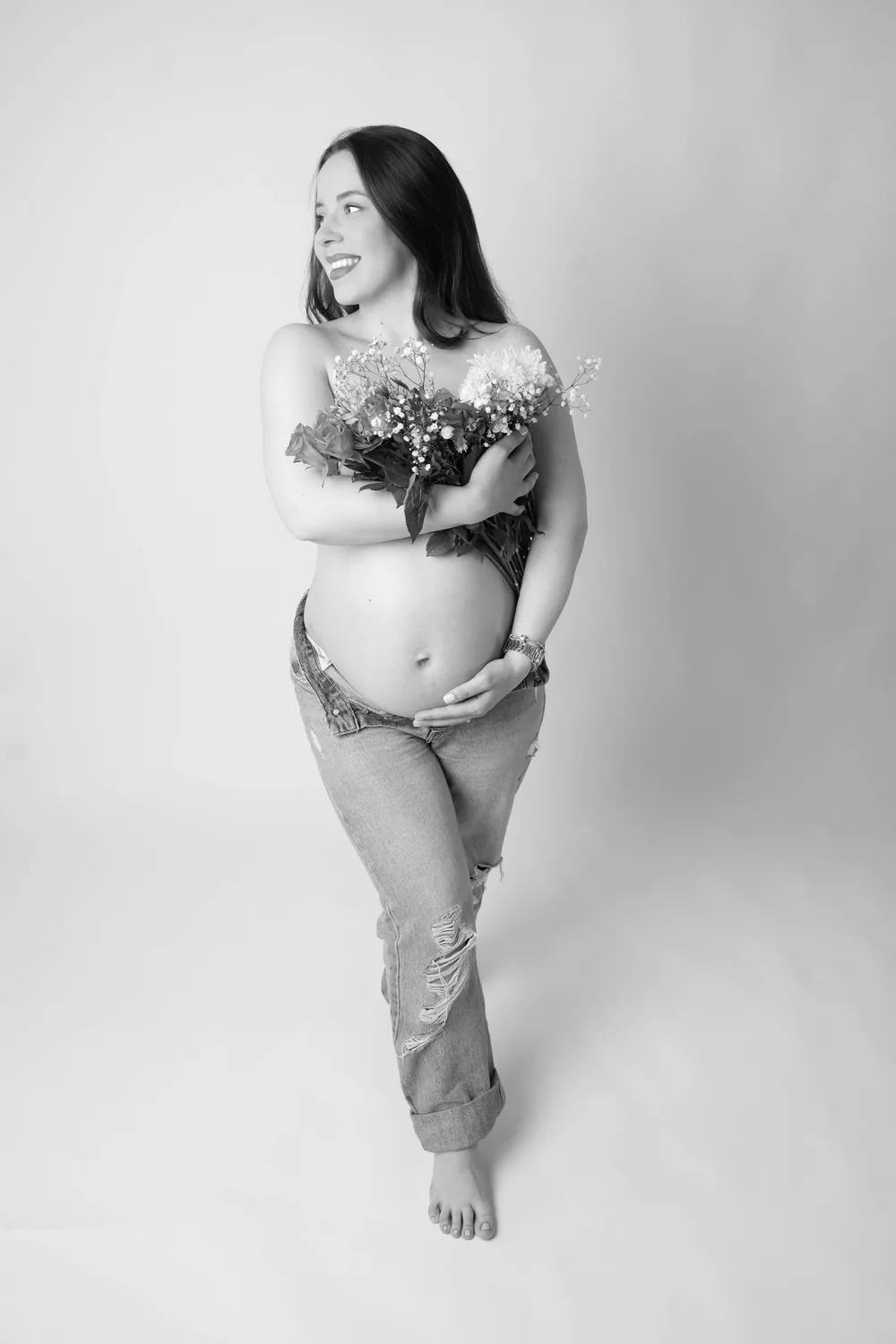 Schwarzweißfoto einer barfüßigen Schwangeren in Jeans, die mit Blumen vor der Brust zur Seite lacht
