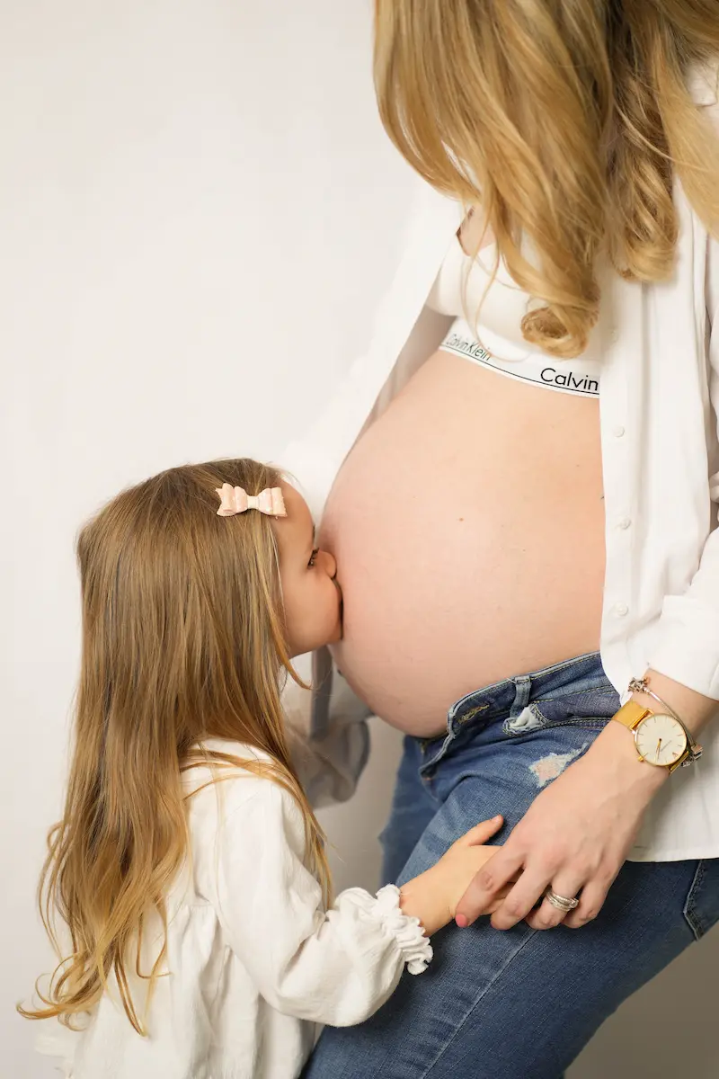 Kind mit Schleife im Haar küsst den Babybauch einer Frau in offenem weißem Hemd