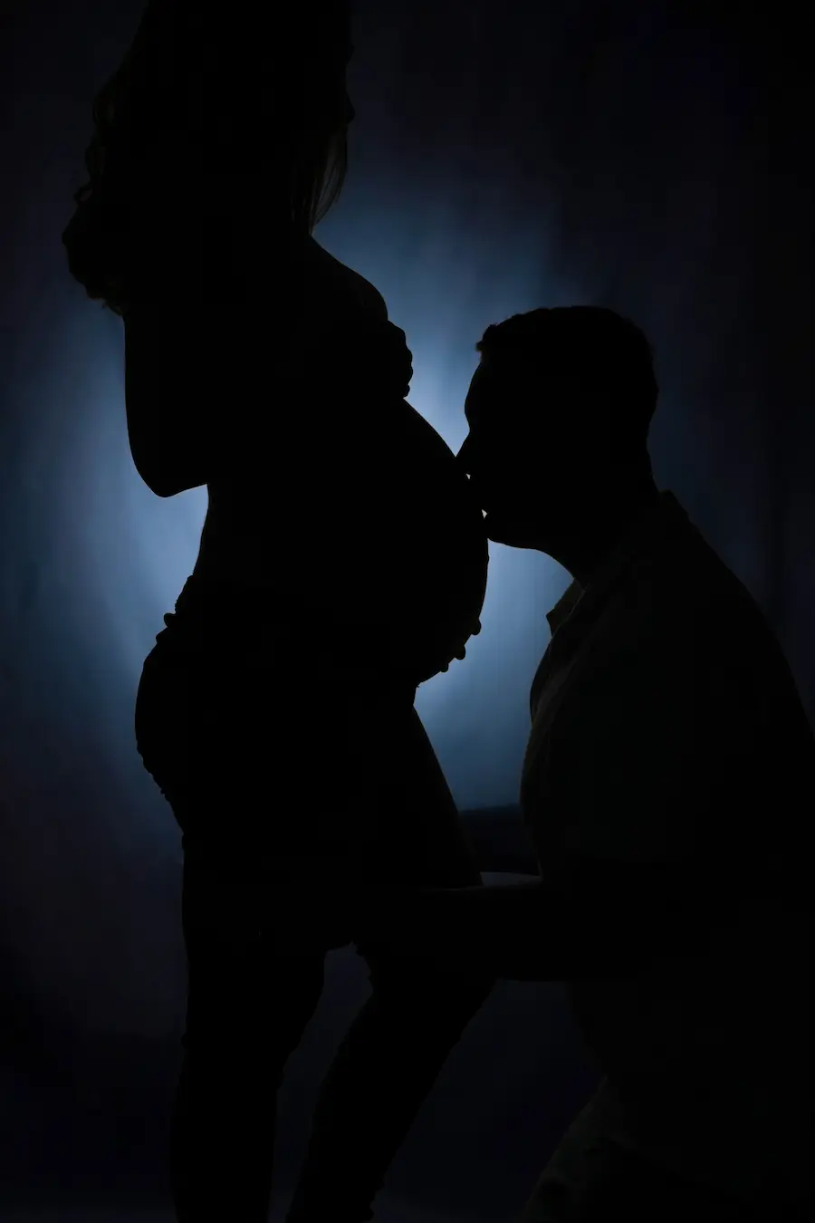 Dunkle Silhouette eines knienden Mannes, der den Babybauch küsst, vor blauem Lichtschein