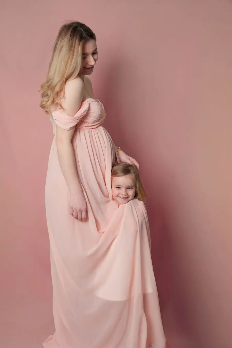 Ein Mädchen schaut unter dem langen rosa Kleid einer schwangeren Frau hervor