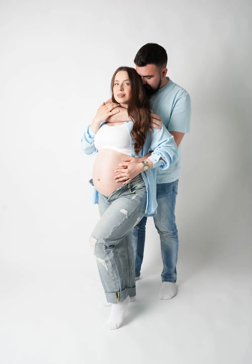 Schwangere Frau im offenen hellblauen Hemd lehnt sich in die Umarmung eines Mannes im blauen Poloshirt