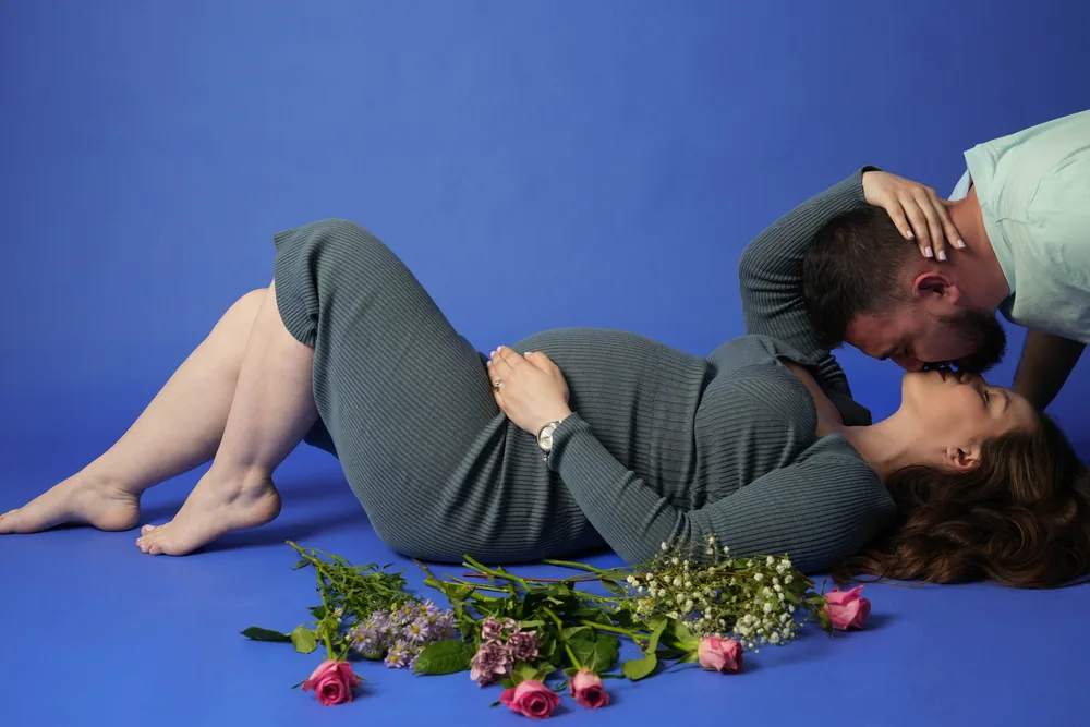 Mann beugt sich zum Kuss über eine schwangere Frau im grauen Kleid, die neben Blumen auf blauem Boden liegt
