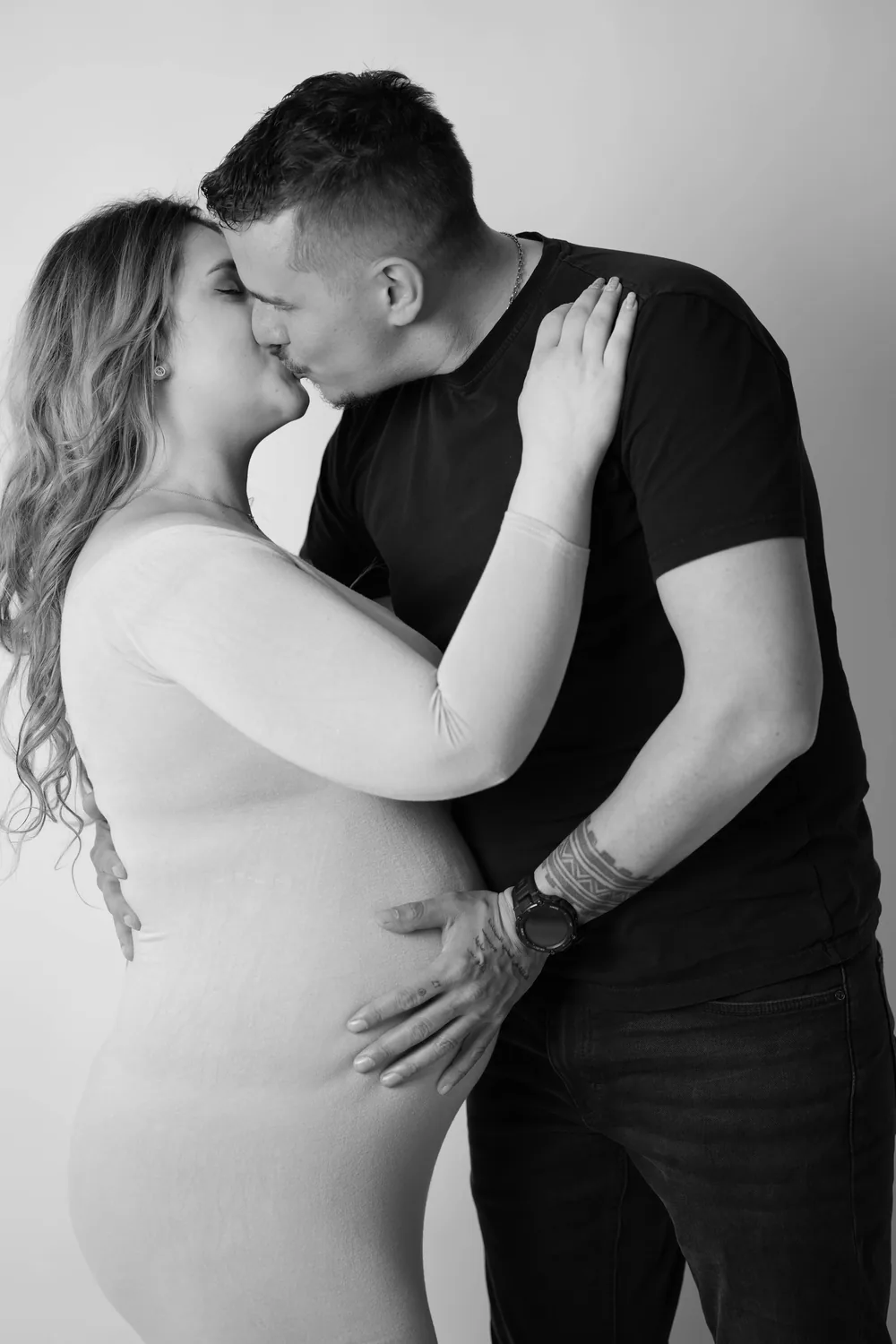 Schwarzweißes Babybauchfoto eines küssenden Paares; die Frau legt einen Arm um den Hals des Mannes