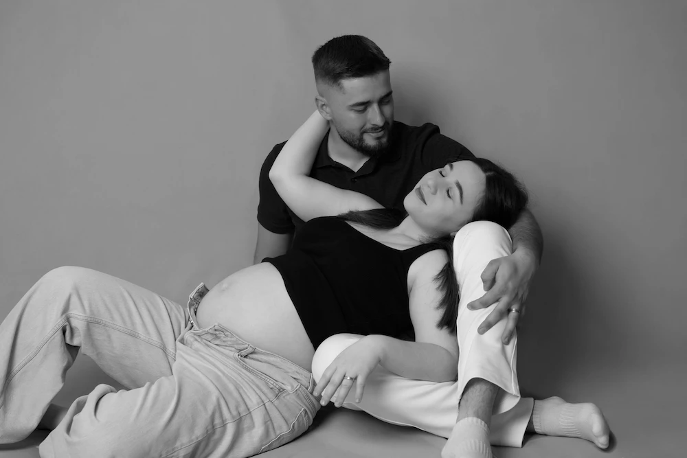 Schwarzweißfoto eines sitzenden Mannes, der eine schwangere Frau seitlich in seinen Armen hält