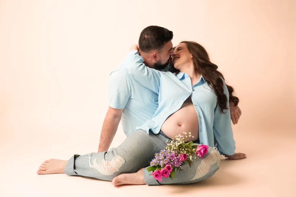 Paar in hellblauen Oberteilen küsst sich im Sitzen auf dem Boden; ein Blumenstrauß liegt unterhalb des Babybauchs
