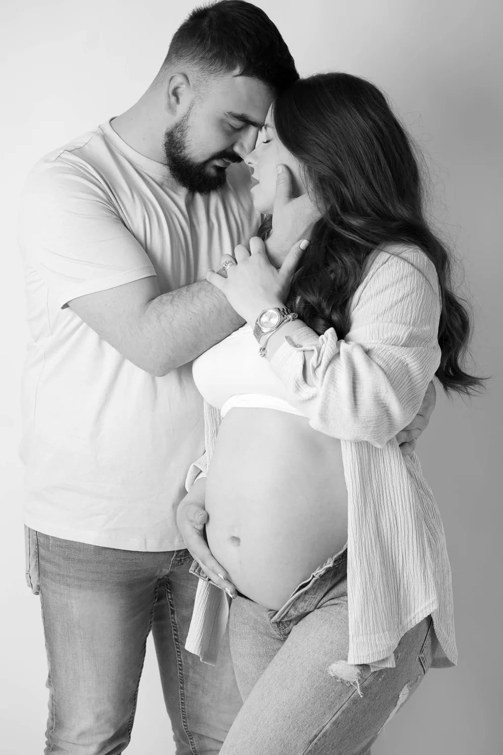 Schwarzweißporträt eines Paares mit aneinandergelegten Stirnen und gemeinsam gehaltenen Händen neben dem Babybauch