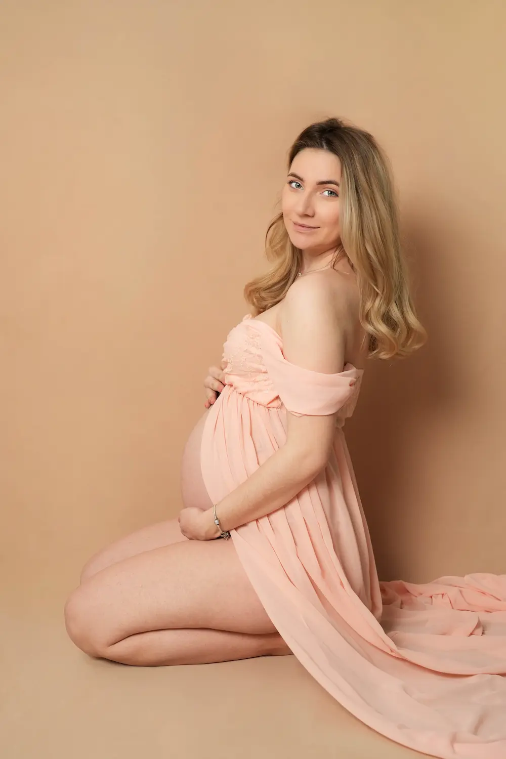 Babybauchfoto einer knienden Frau im roséfarbenen Kleid mit langer Schleppe vor beigem Hintergrund