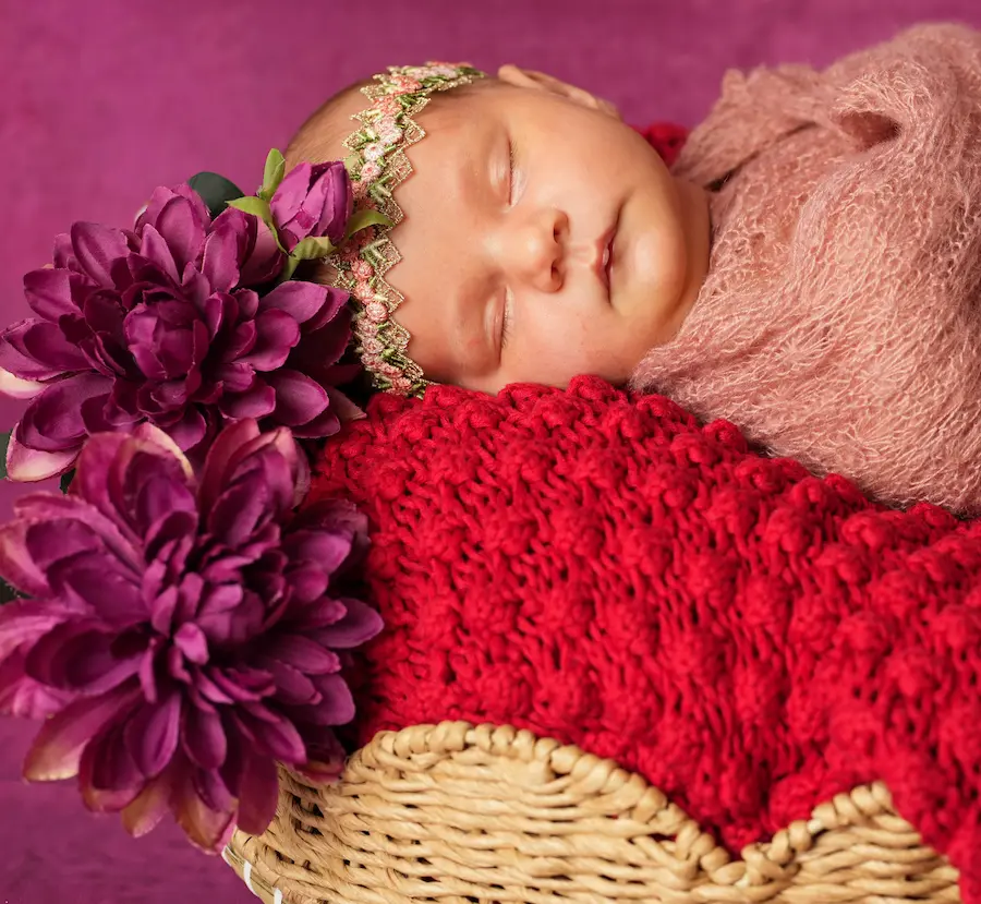 Schlafendes Babygesicht mit feinem Blumenband neben violetten Blüten und einer roten Strickdecke