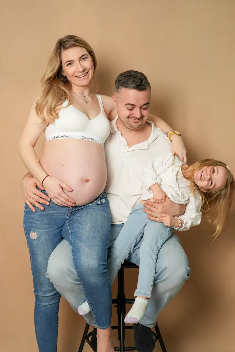 Schwangere Frau neben einem sitzenden Mann, der ein lachendes Kind auf dem Arm hält