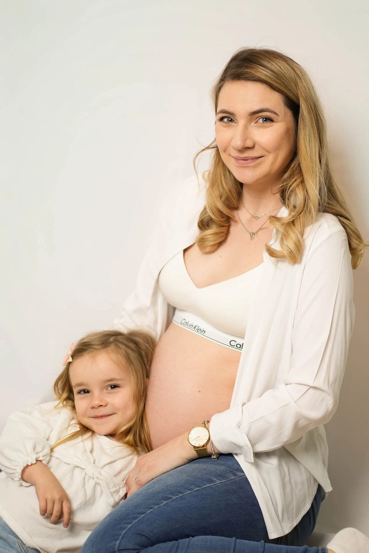 Schwangere Frau im weißen Hemd sitzt neben einem lächelnden Kind, das sich an ihren Babybauch schmiegt