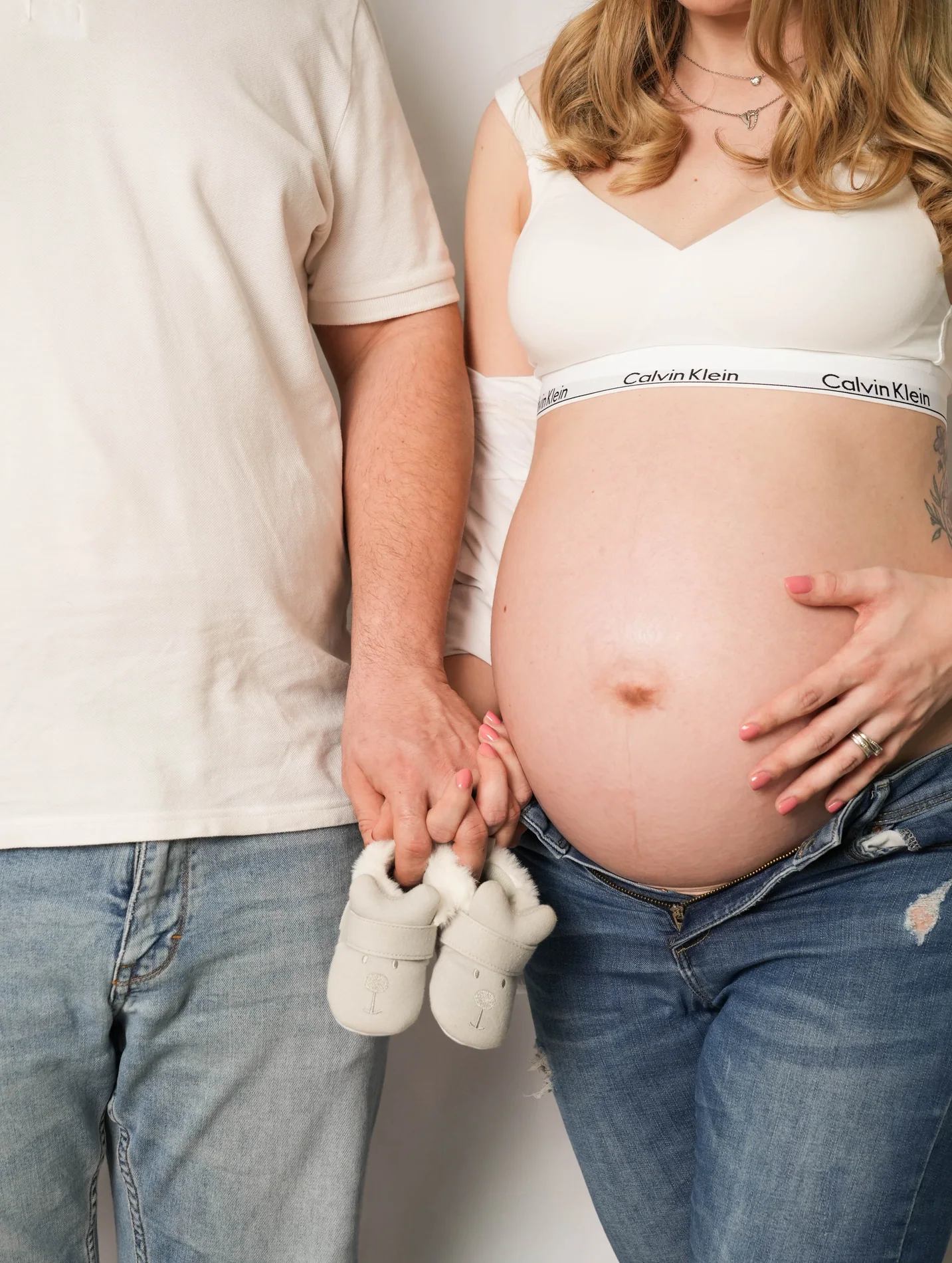 Farbiges Bauchdetail eines Paares in weißen Oberteilen und Jeans mit hellen Babyschuhen in den Händen