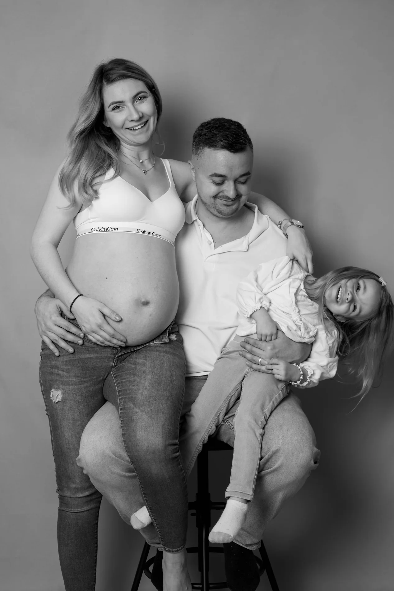 Schwarzweißfoto einer Schwangeren neben einem sitzenden Mann, der ein lachendes Kind auf dem Schoß hält