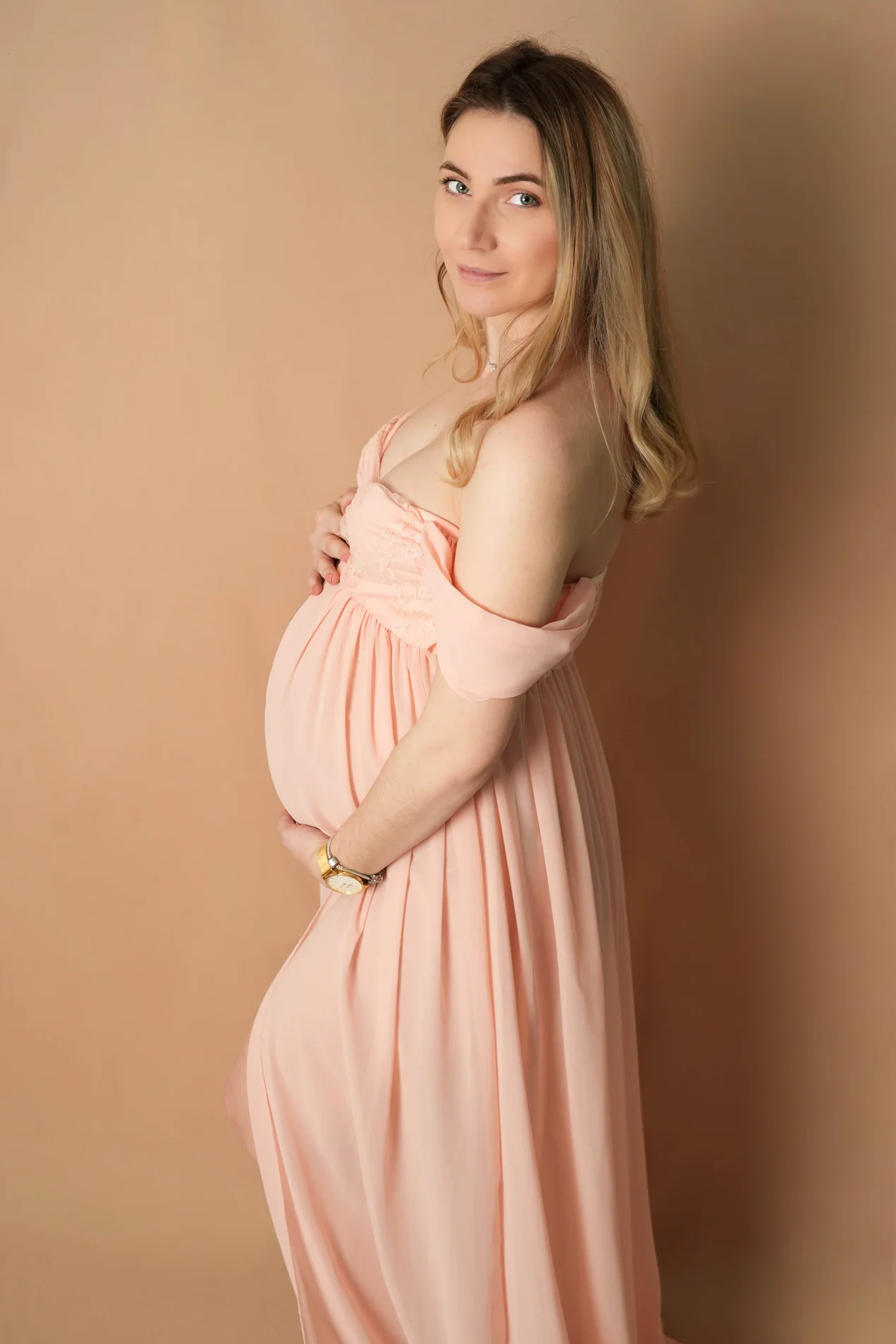 Schwangerschaftsporträt im roséfarbenen Kleid mit Blick in die Kamera und beiden Händen am Bauch