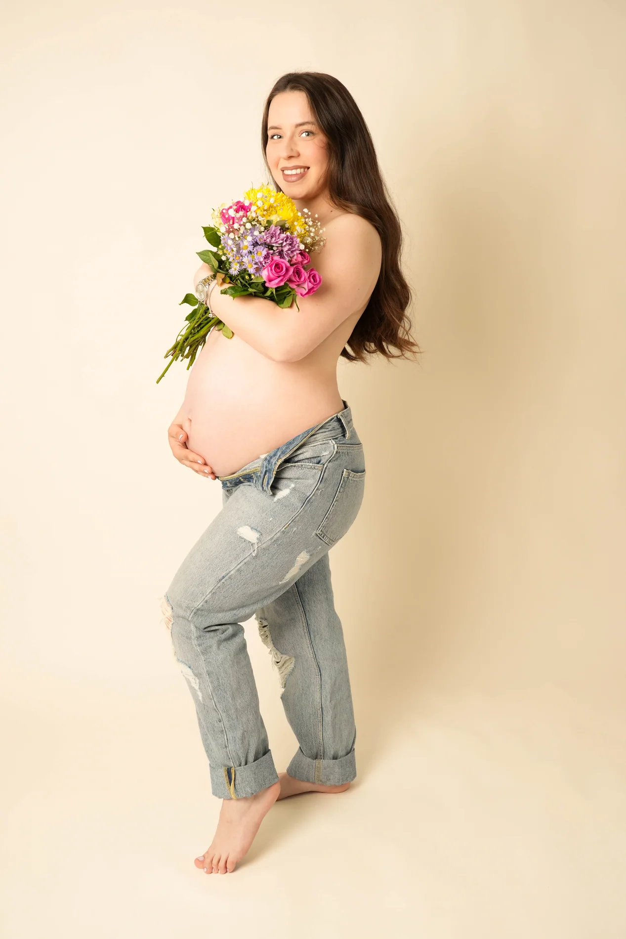 Seitliches Babybauchfoto einer lächelnden Frau in Jeans mit buntem Blumenstrauß vor beigem Hintergrund
