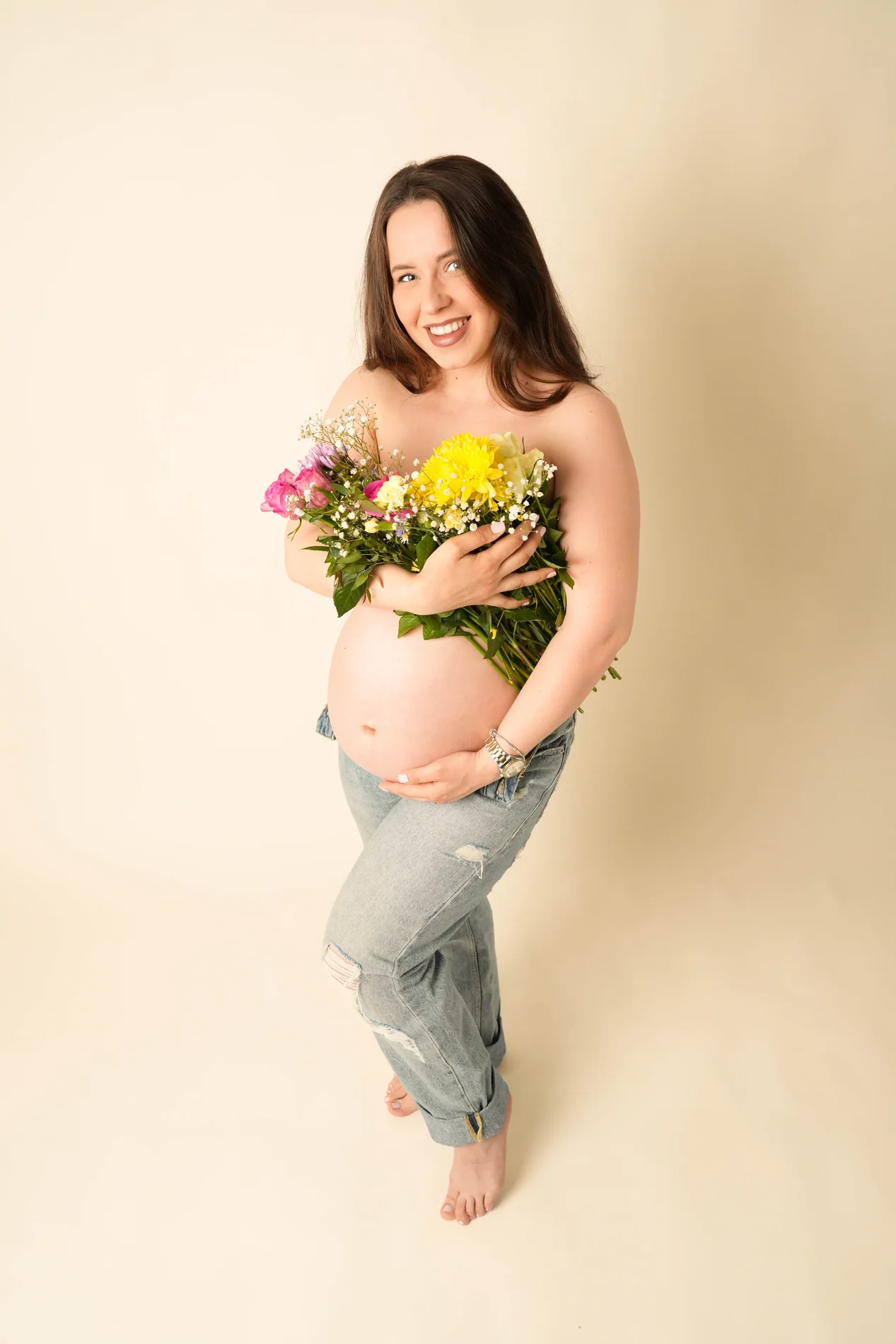 Barfüßige Schwangere in Jeans hält einen bunten Blumenstrauß vor der Brust