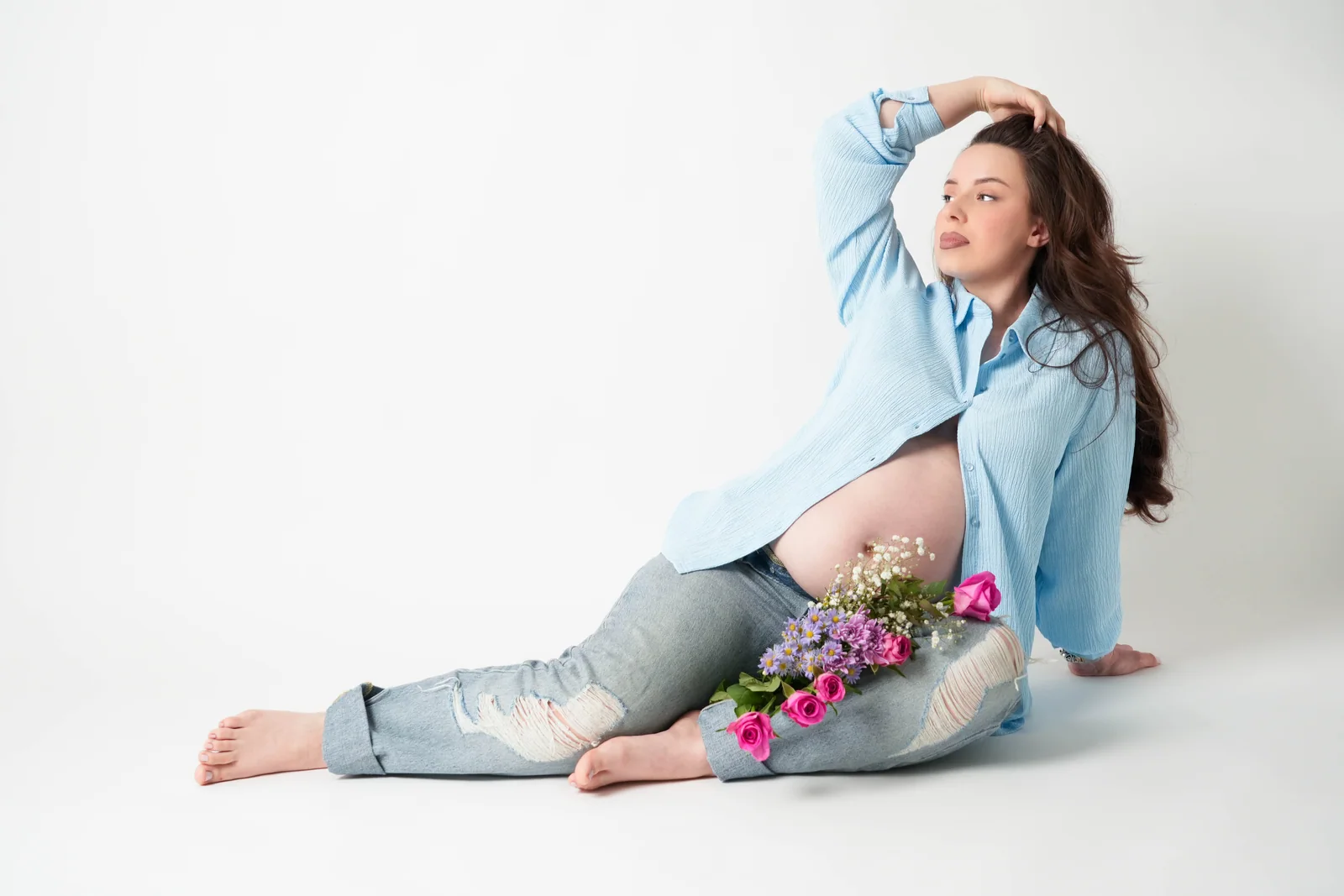 Schwangere Frau im blauen Hemd sitzt mit einer Hand im Haar auf dem Boden, mit rosa Blumen auf dem Schoß