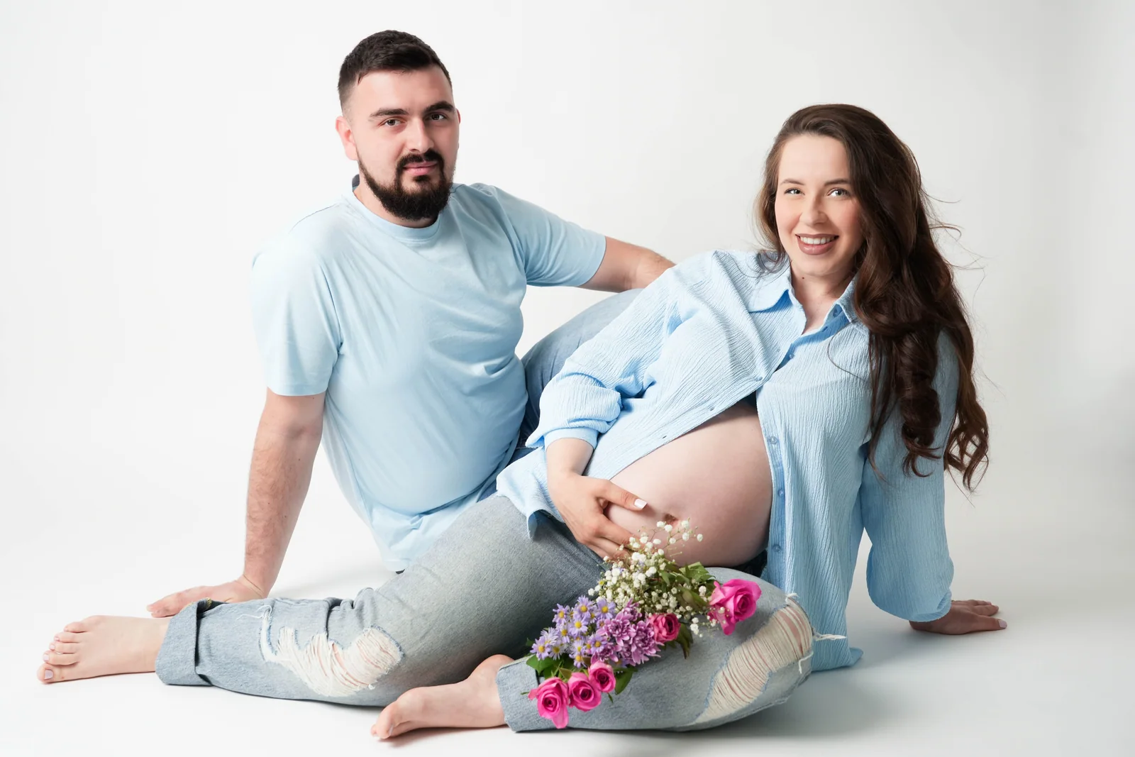 Sitzendes Paar in Hellblau beim Babybauch-Shooting; die Frau lacht mit Blumen auf den ausgestreckten Beinen