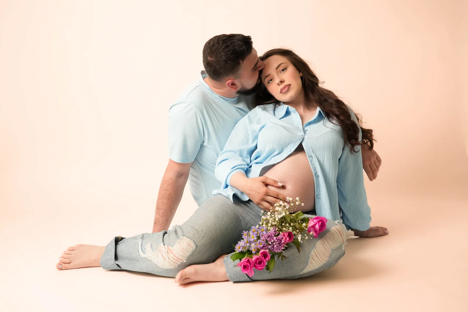 Mann küsst die Schläfe einer schwangeren Frau; beide sitzen in blauer Kleidung neben einem rosa Blumenstrauß