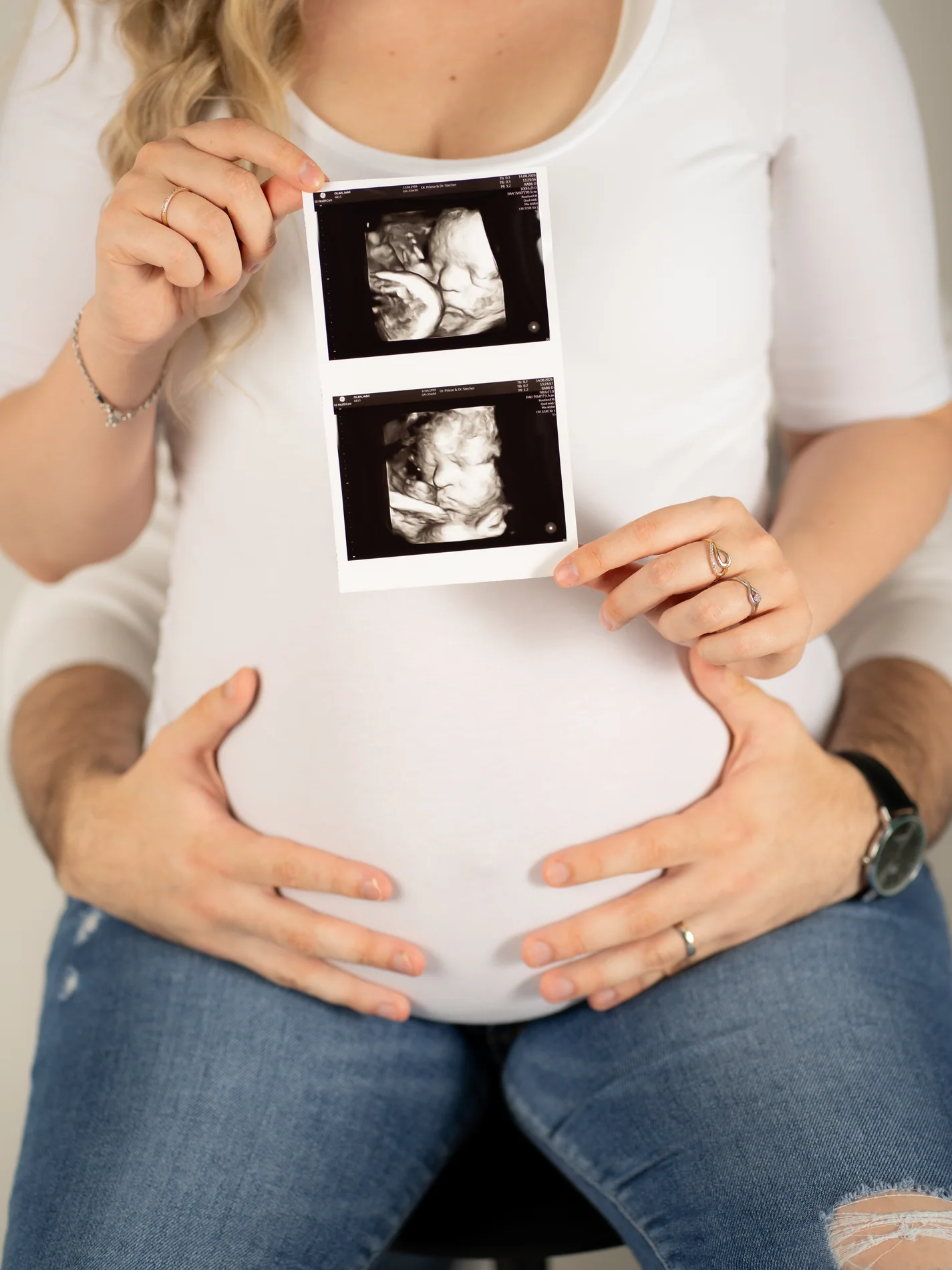 Nahaufnahme eines Babybauchs im weißen Oberteil mit zwei Ultraschallbildern darüber und Händen an der Bauchunterseite