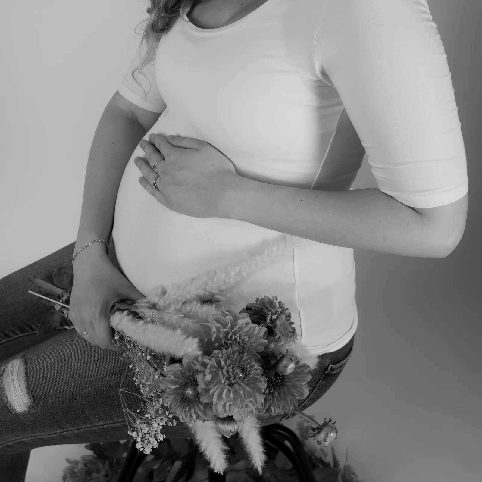 Schwarzweißdetail von Händen am Babybauch unter einem weißen Oberteil, mit Blumen im Vordergrund