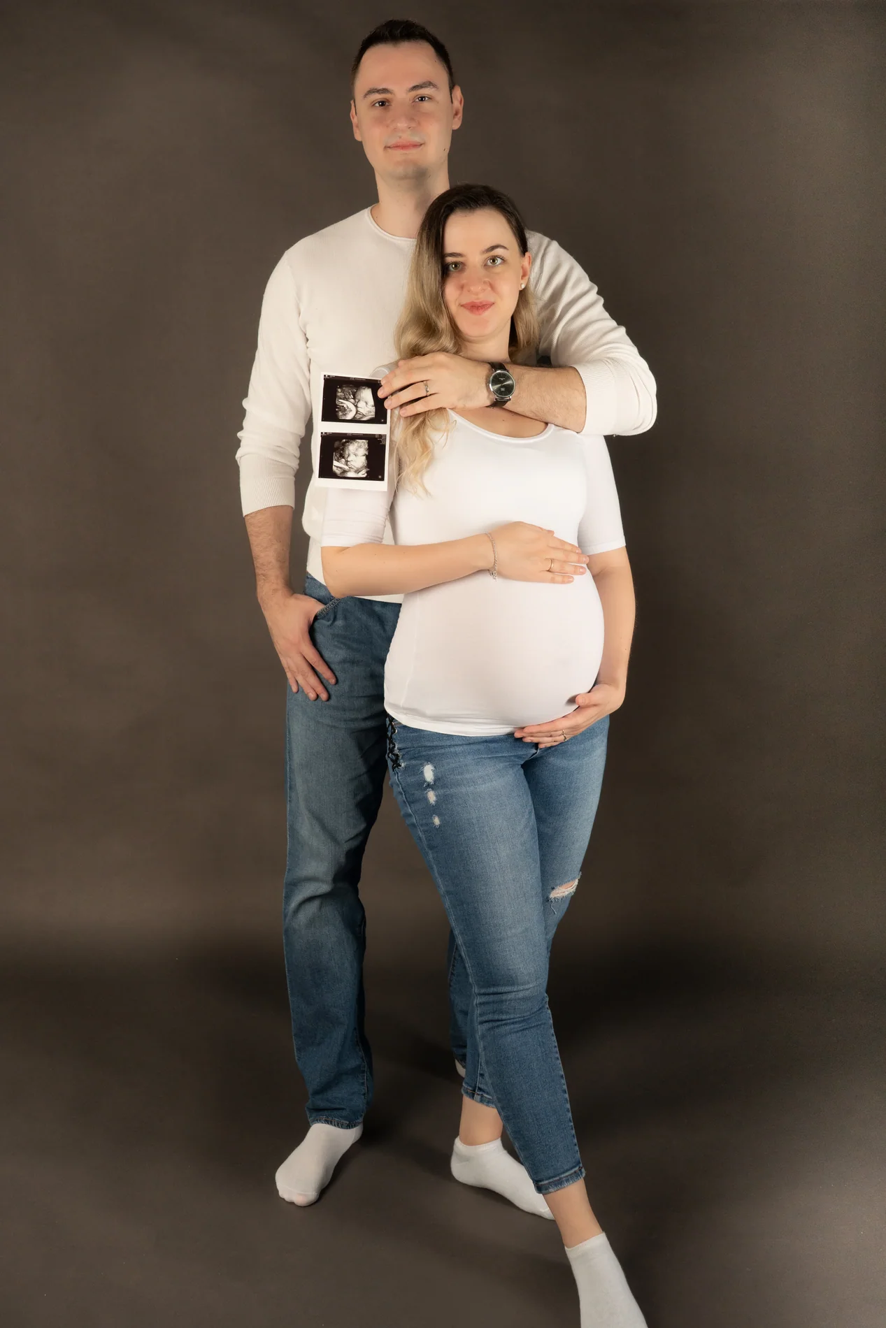 Ganzkörperfoto eines Paares mit Ultraschallbildern vor graubraunem Hintergrund; beide tragen Jeans und weiße Socken