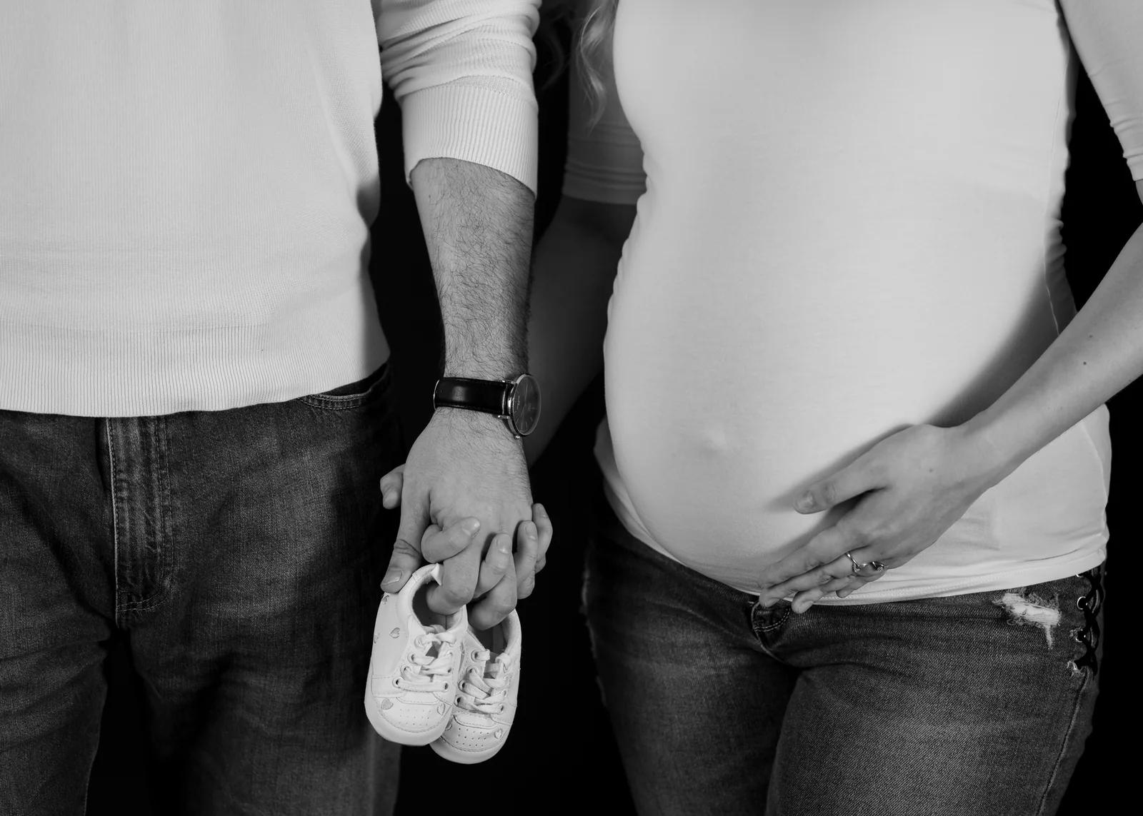 Schwarzweißdetail eines Paares in Jeans: verschränkte Hände mit Babyschuhen neben dem Babybauch