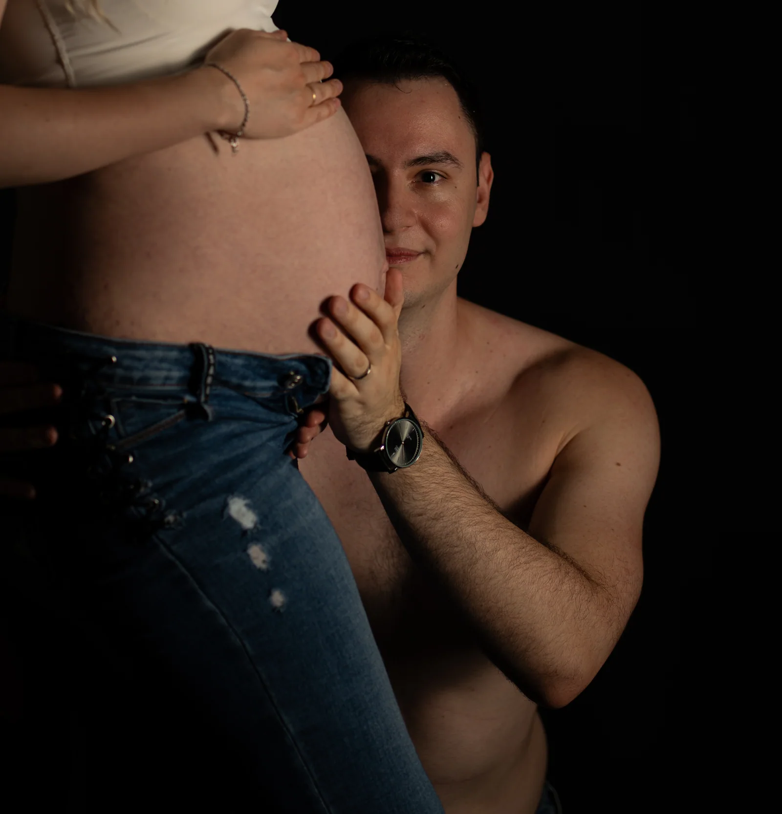 Mann legt das Gesicht seitlich an den Babybauch und blickt vor dunklem Hintergrund in die Kamera
