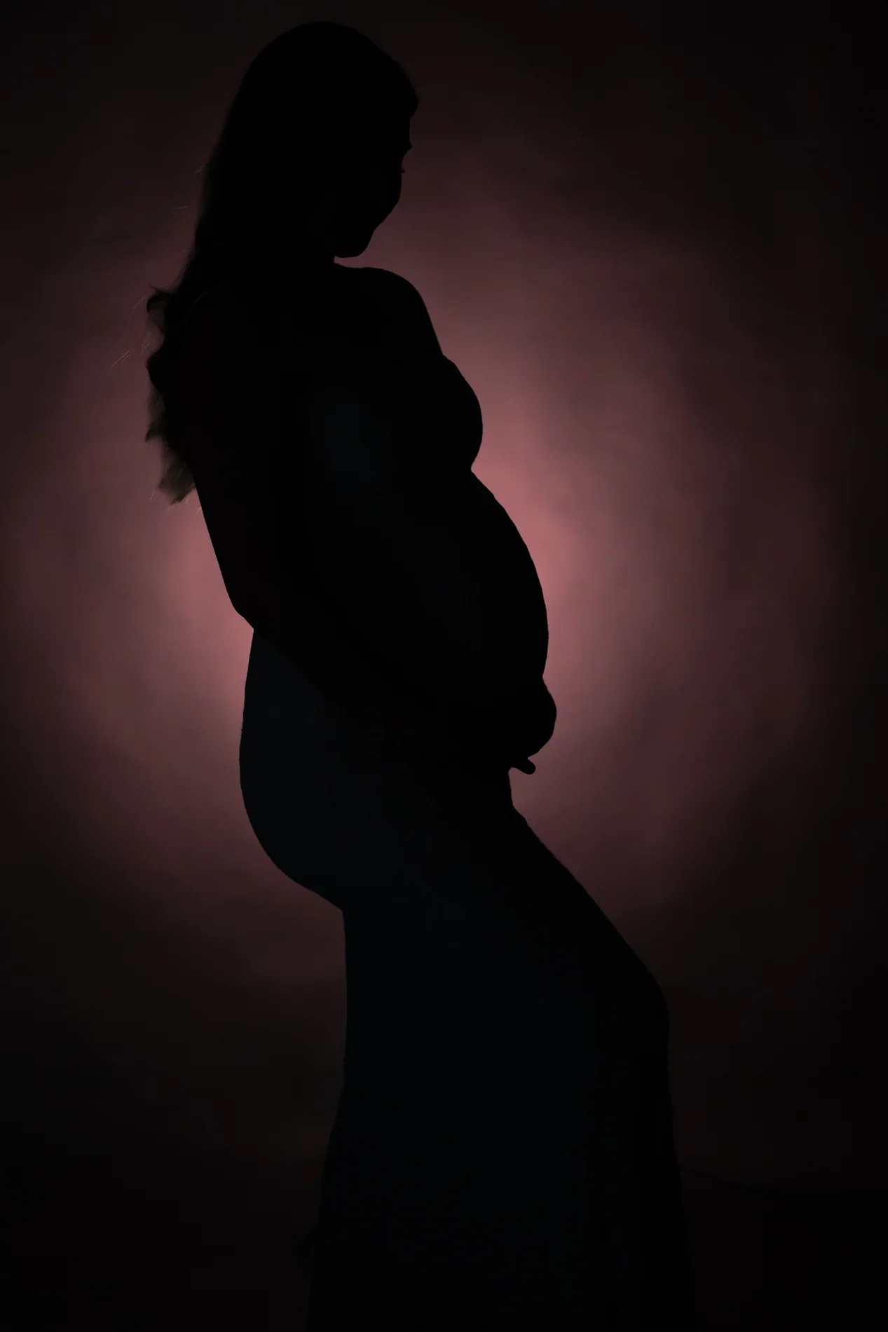 Seitliche Silhouette einer Schwangeren mit Händen unter dem Bauch vor rosa Lichtschein