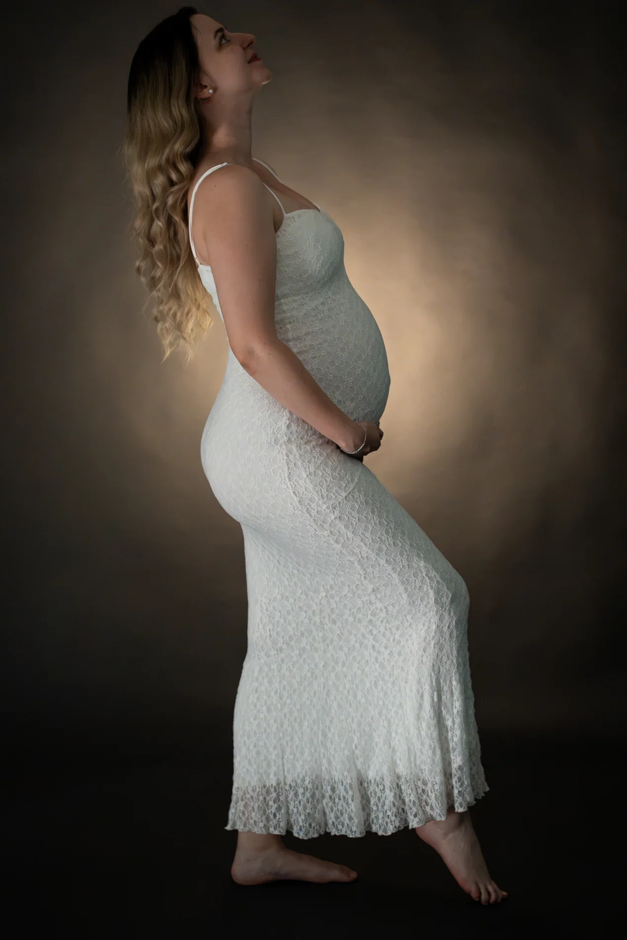 Seitliches Schwangerschaftsporträt im weißen Spitzenkleid mit nach oben gerichtetem Blick vor dunklem Hintergrund