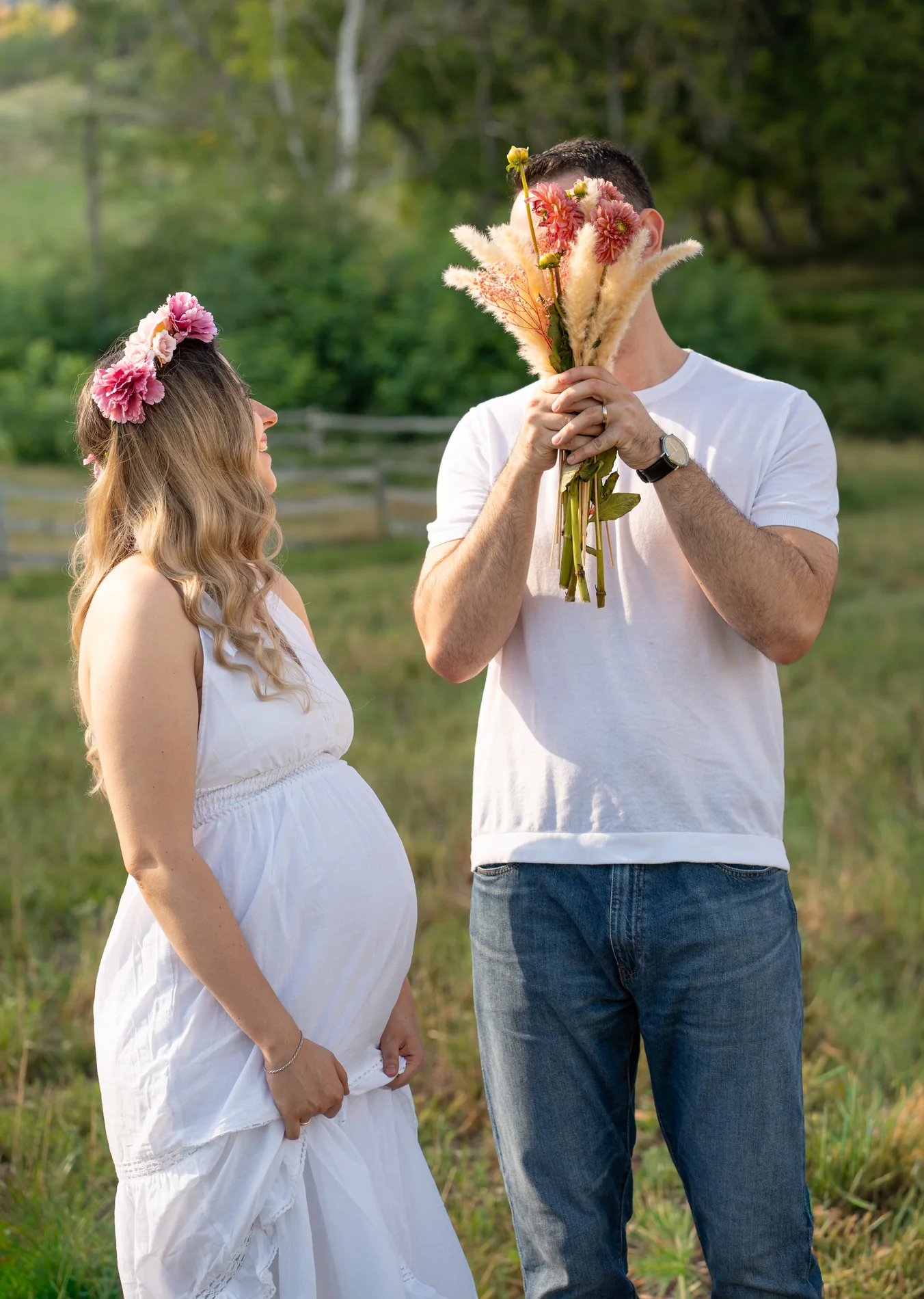Babybauchfoto im Grünen: Eine Frau mit Blumenkranz schaut zu einem Mann, der sein Gesicht hinter einem Strauß versteckt