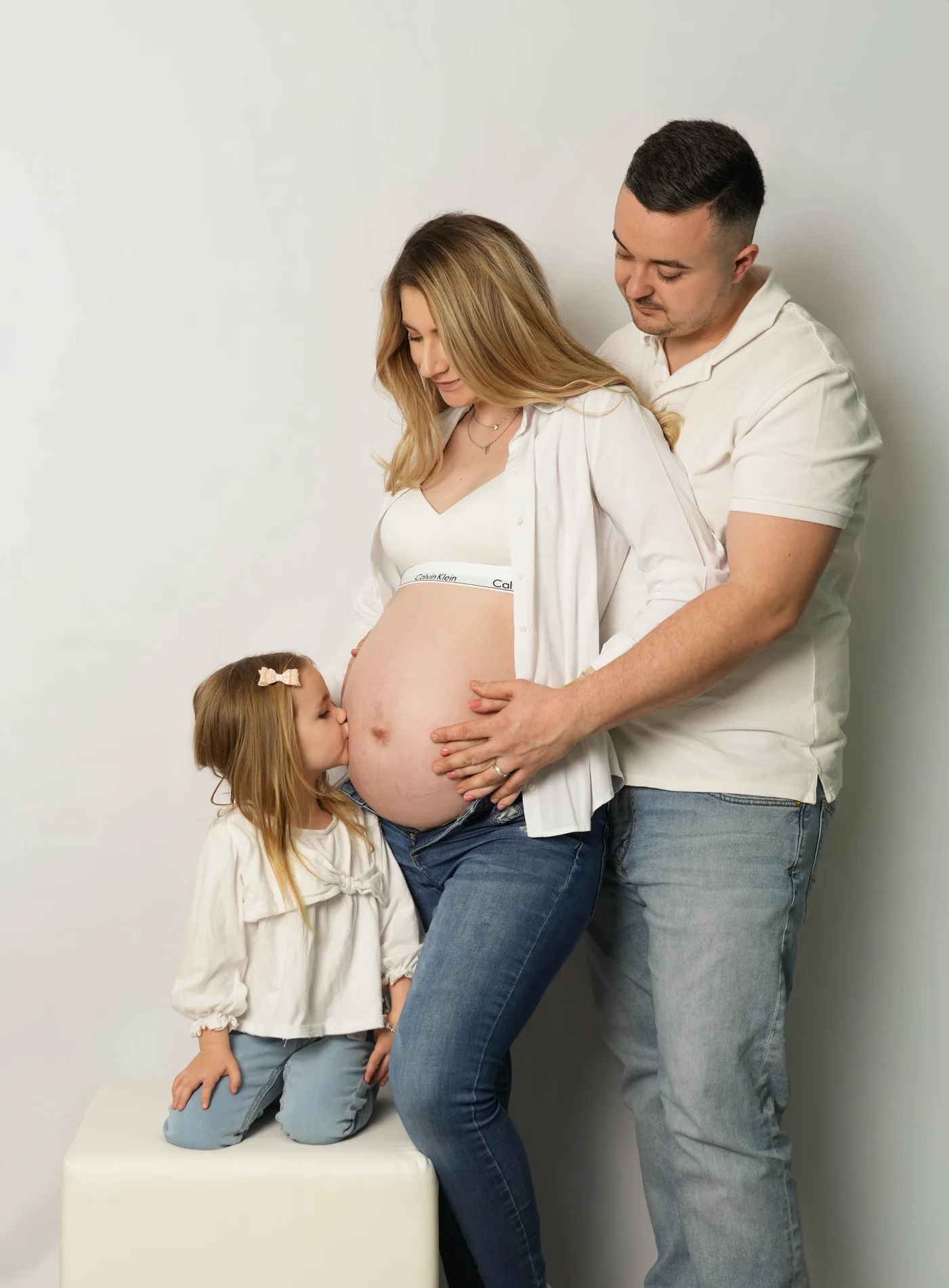 Ein Mädchen küsst einen Babybauch, während ein Mann die schwangere Frau von hinten hält