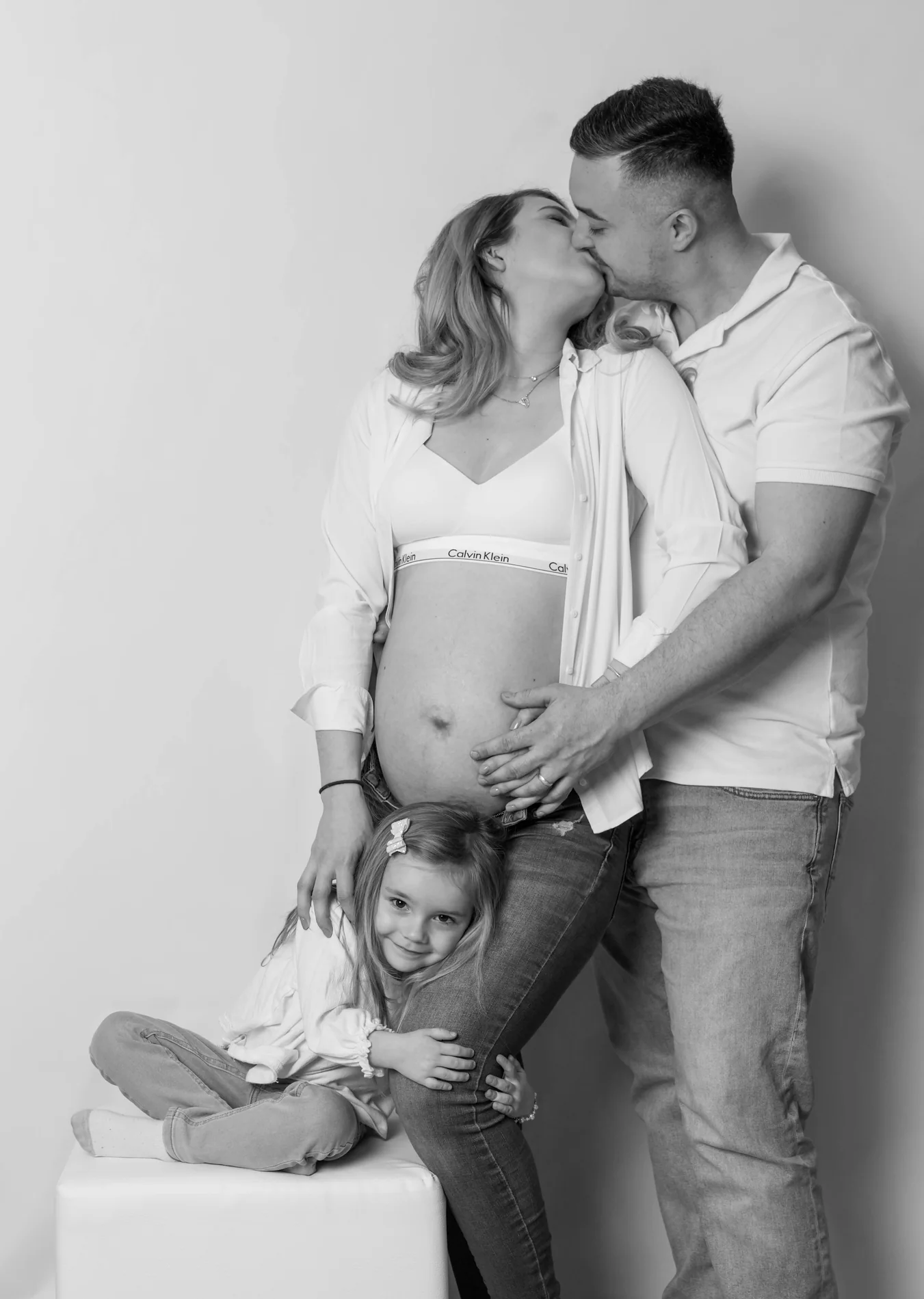 Schwarz-Weiß-Familienfoto: Ein Paar küsst sich, ein Mädchen sitzt neben dem Babybauch