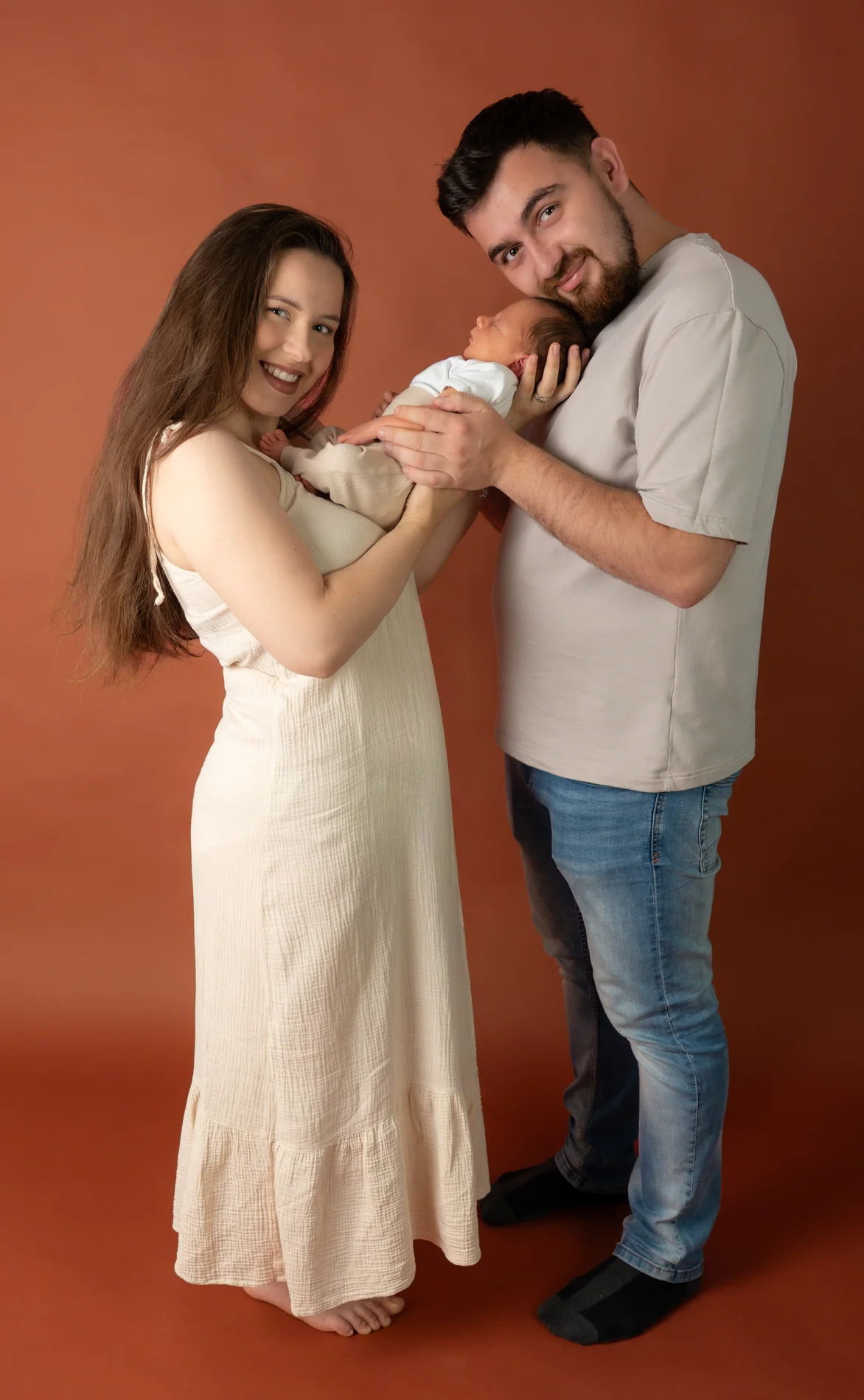 Ein Paar mit Baby im Arm steht vor rostbraunem Hintergrund und lächelt zur Kamera