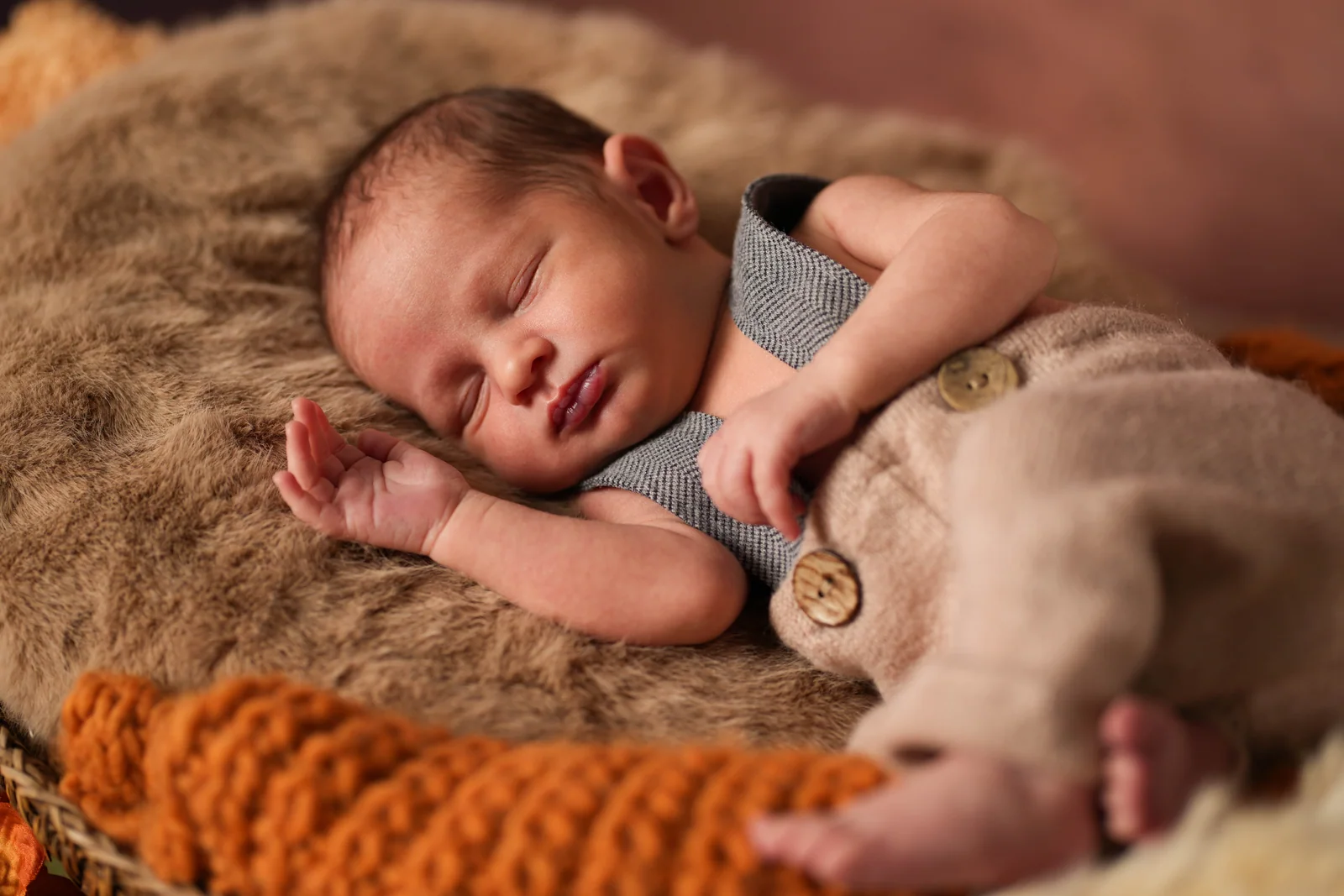 Schlafendes Baby in brauner Hose mit einem angewinkelten Arm neben dem Kopf auf weicher Unterlage