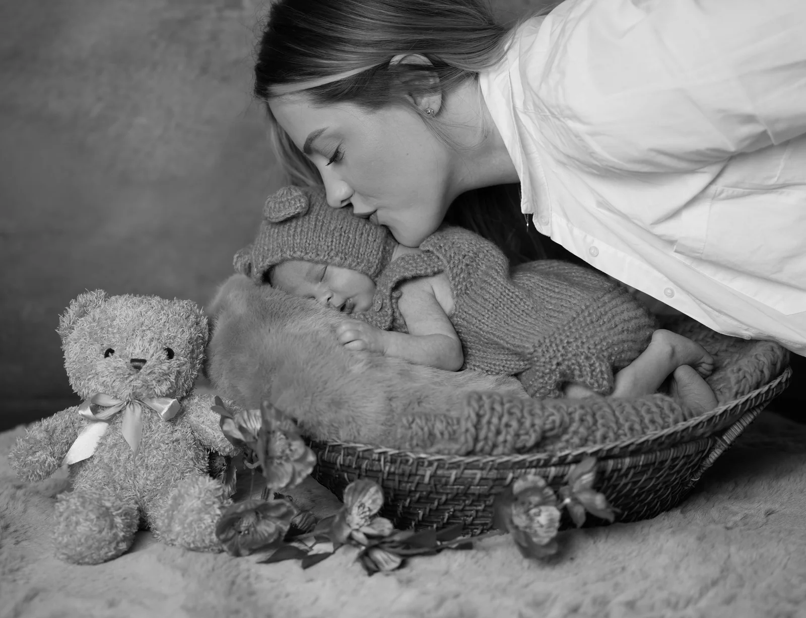 Schwarzweißaufnahme einer Frau, die ein Baby mit Strickmütze im Korb neben einem Teddy küsst