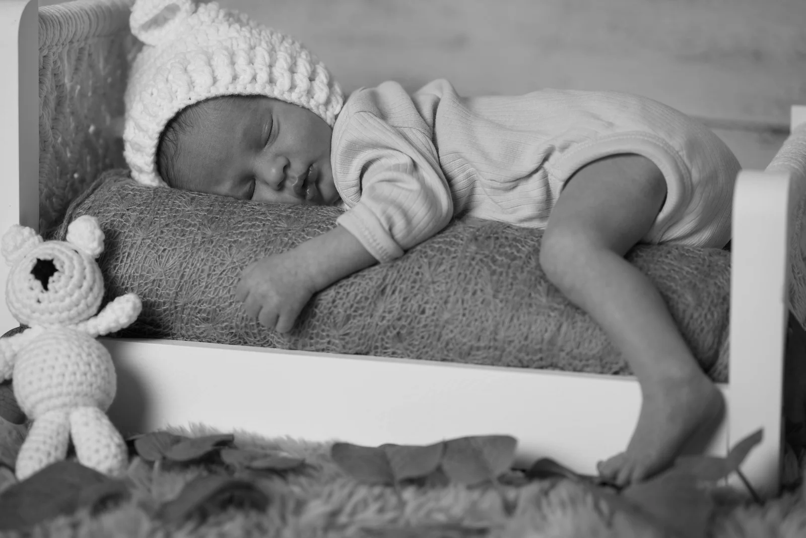 Schwarzweißes Babyfoto im kleinen Holzbett mit einem Bein über dem Bettrand und Häkelbär daneben