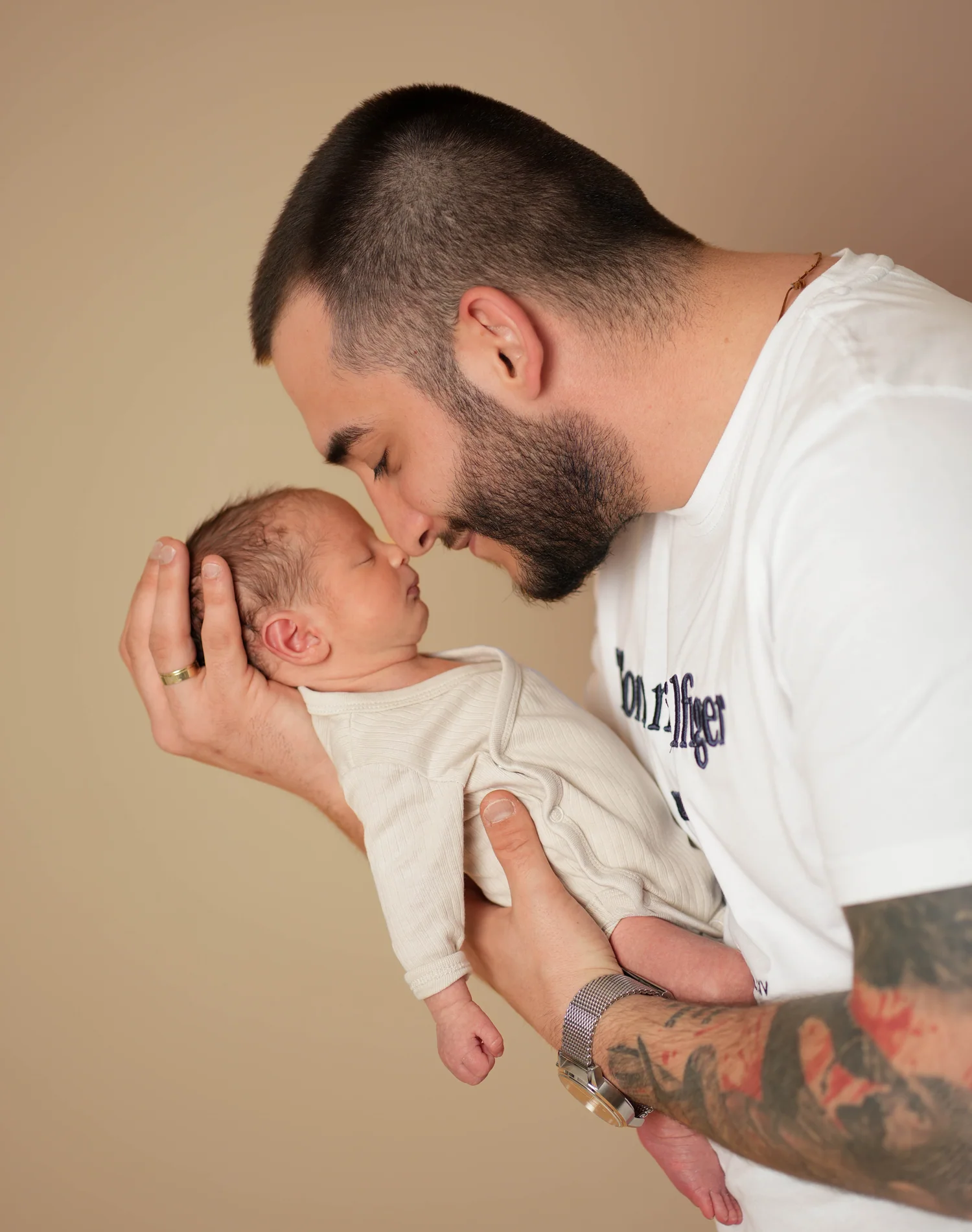 Mann mit tätowiertem Arm hält ein Baby im weißen Strampler nah an sein Gesicht