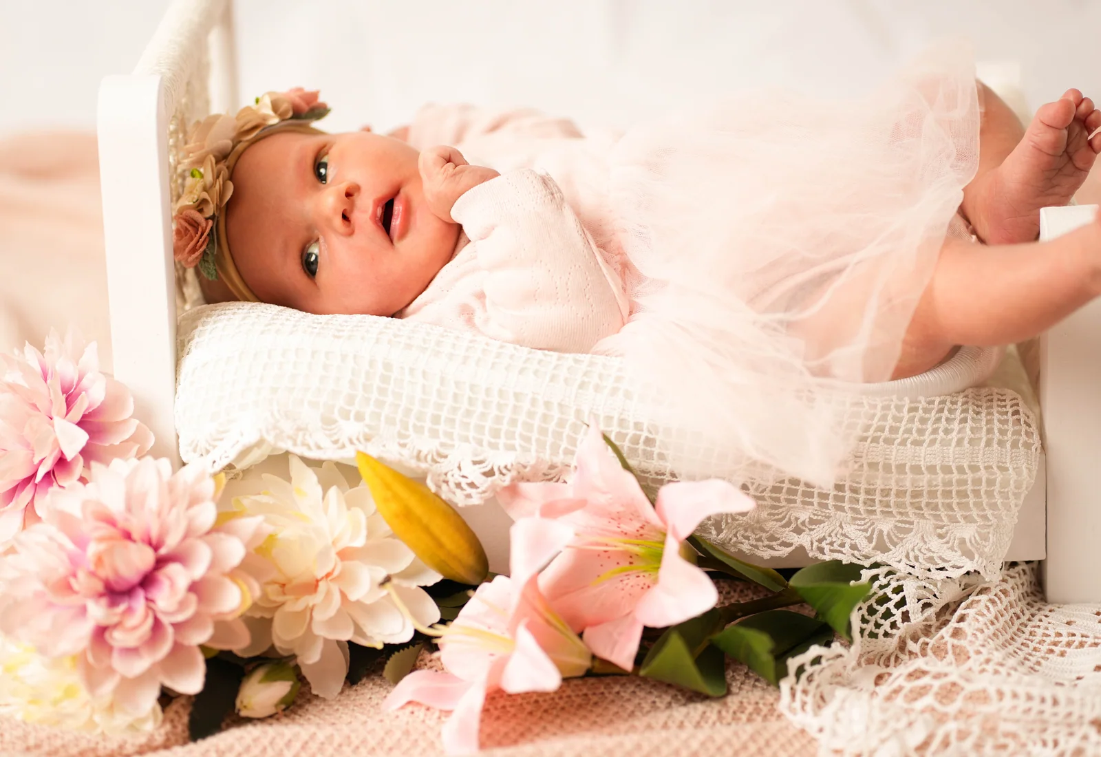 Waches Baby im rosa Spitzenkleid liegt auf einem weißen Kissen neben bunten Blüten