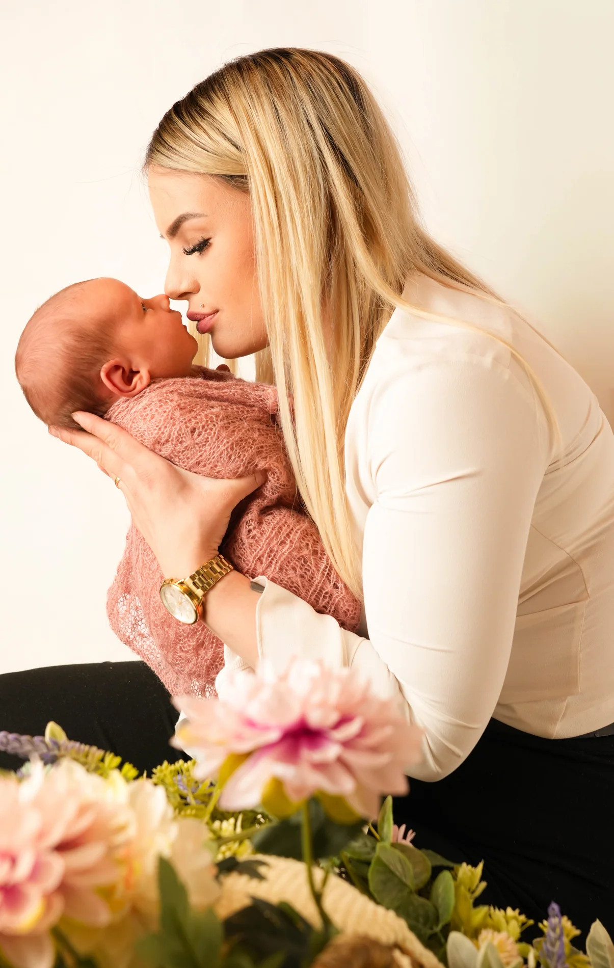 Blonde Frau hält ein rosa eingewickeltes Baby vor sich, Blumen stehen im Vordergrund