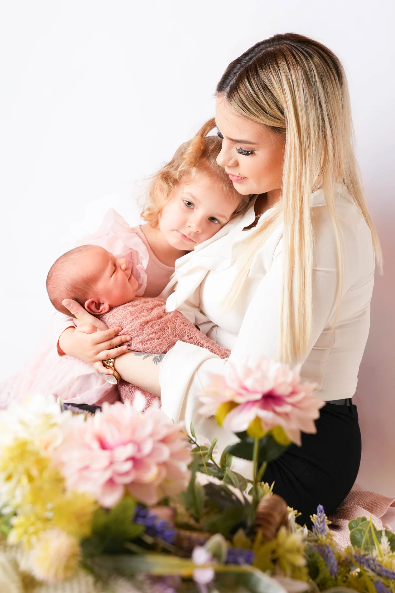 Frau in weißer Bluse hält ein Baby und ein kleines Mädchen eng bei sich, hinter bunten Blumen
